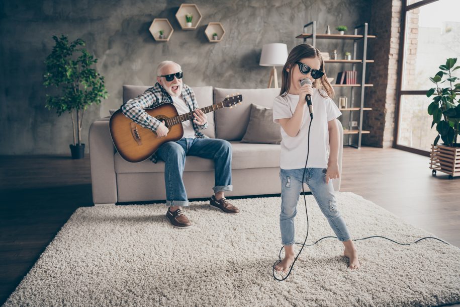 Photo of grandpa play guitar little pretty granddaughter hold mic singing, song spend time rejoicing cool style trendy sun glasses stay home quarantine modern living room indoors
